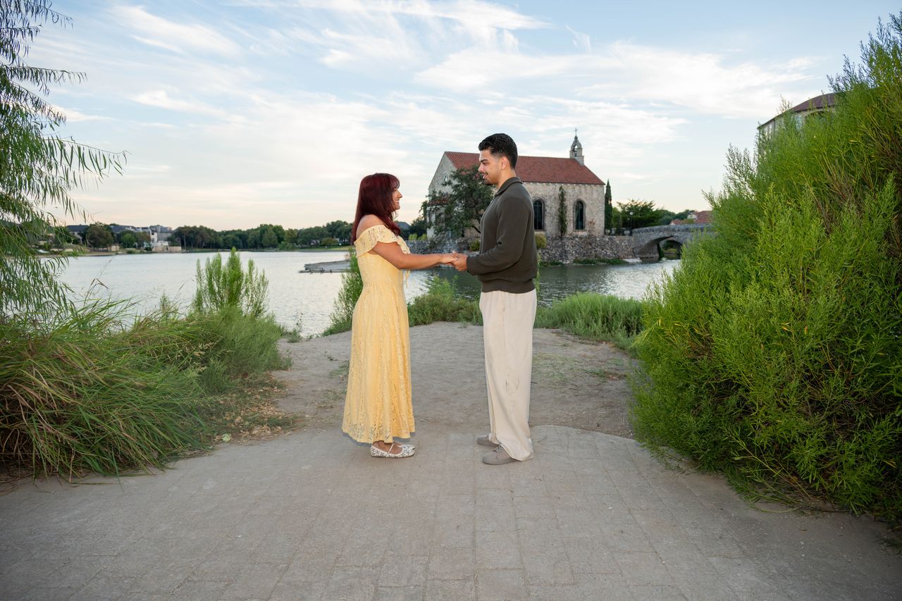 Luca and Ana face each other on the lake path, the chapel behind them