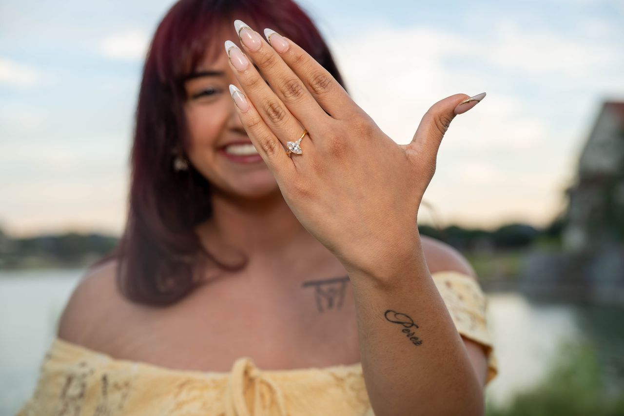 Ana smiles behind her raised hand, showing the engagement ring