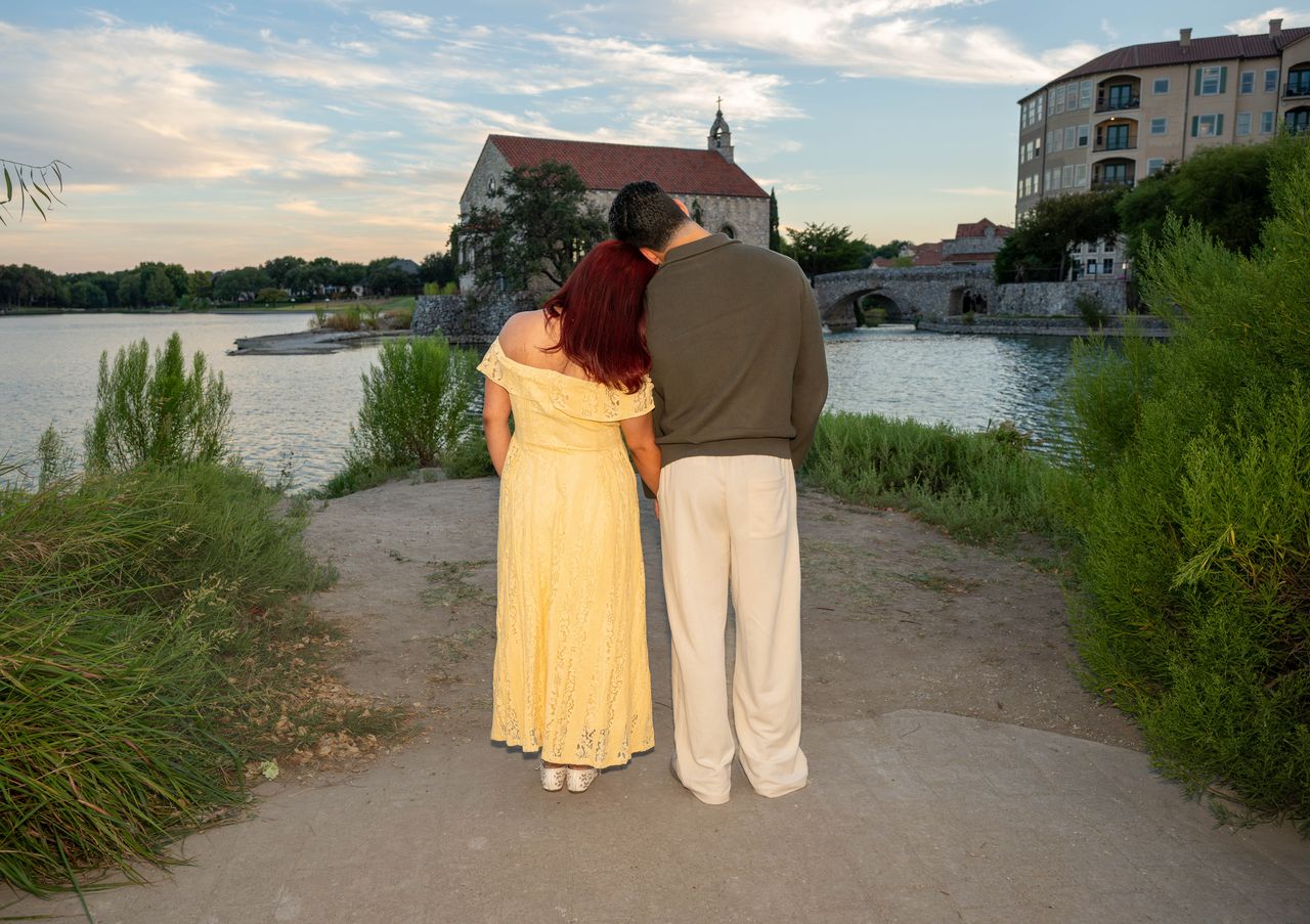 Luca and Ana stand together on the path, facing the lake and the chapel