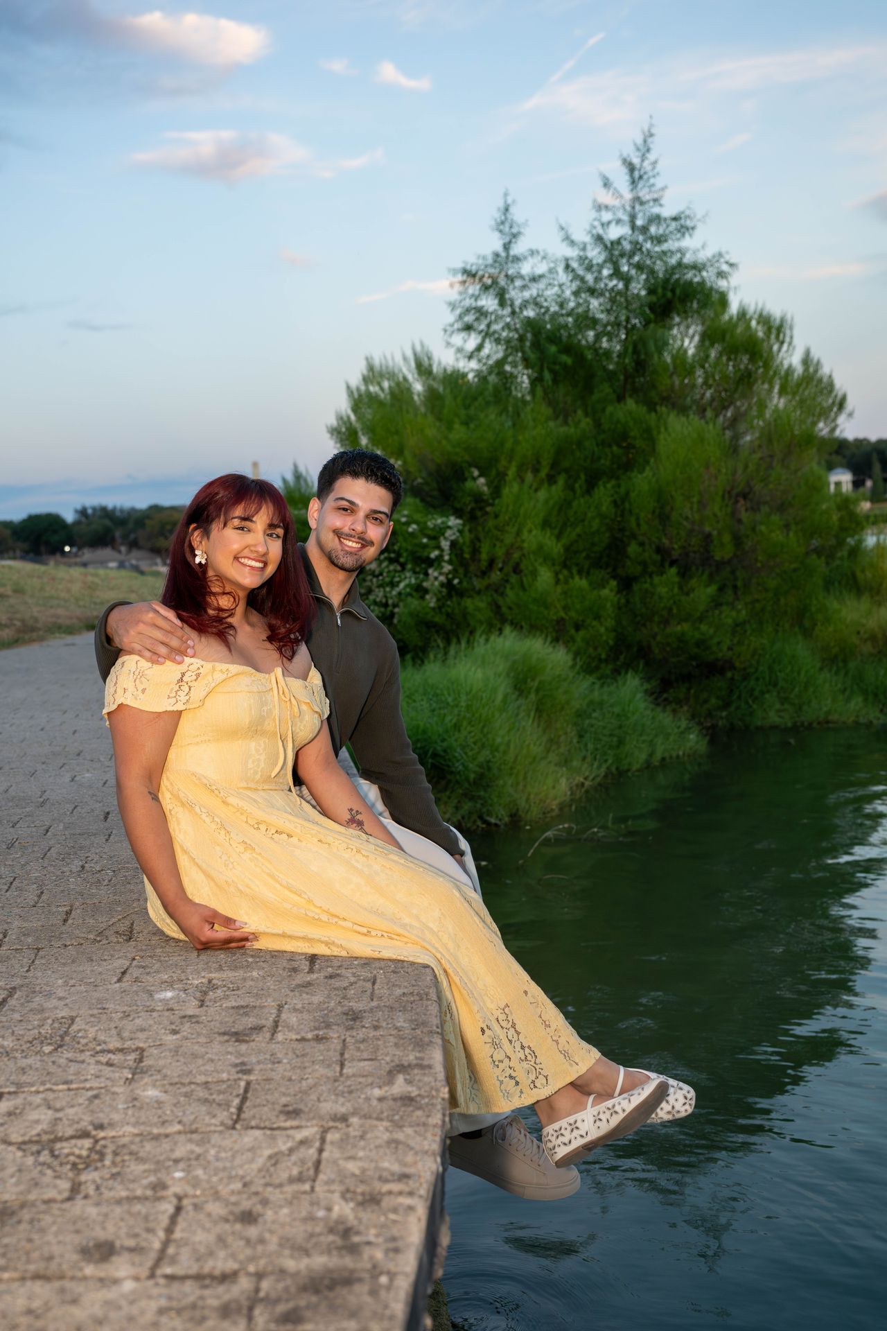 Ana and Luca sit on a stone ledge over the water and smile at the camera