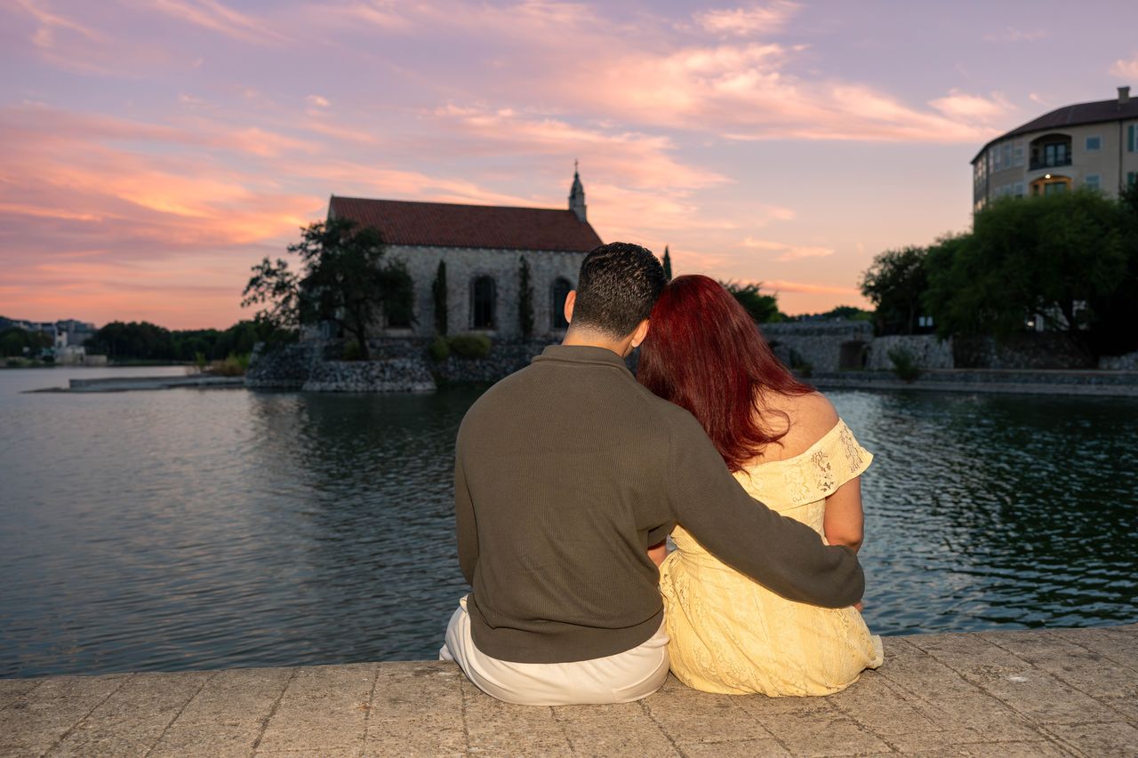 Luca and Ana sit together by the water at sunset, the chapel across the lake