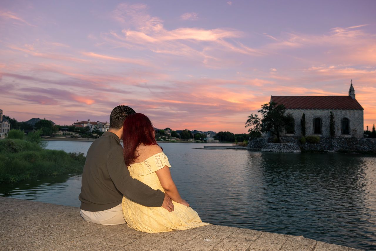 Luca and Ana watch a pink sunset over the lake beside the chapel