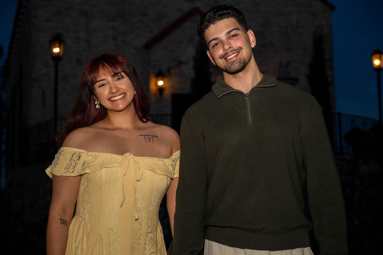 Ana and Luca smile at the camera in front of the chapel lamps