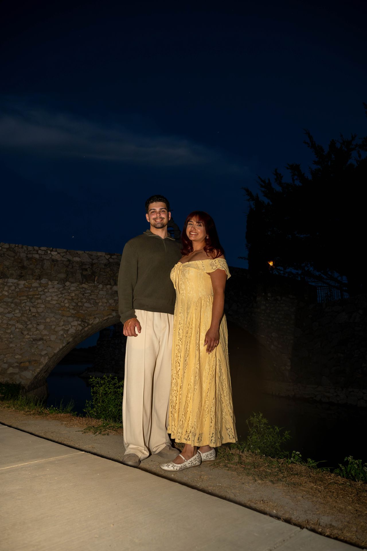 Luca and Ana stand on the path by the stone bridge at night