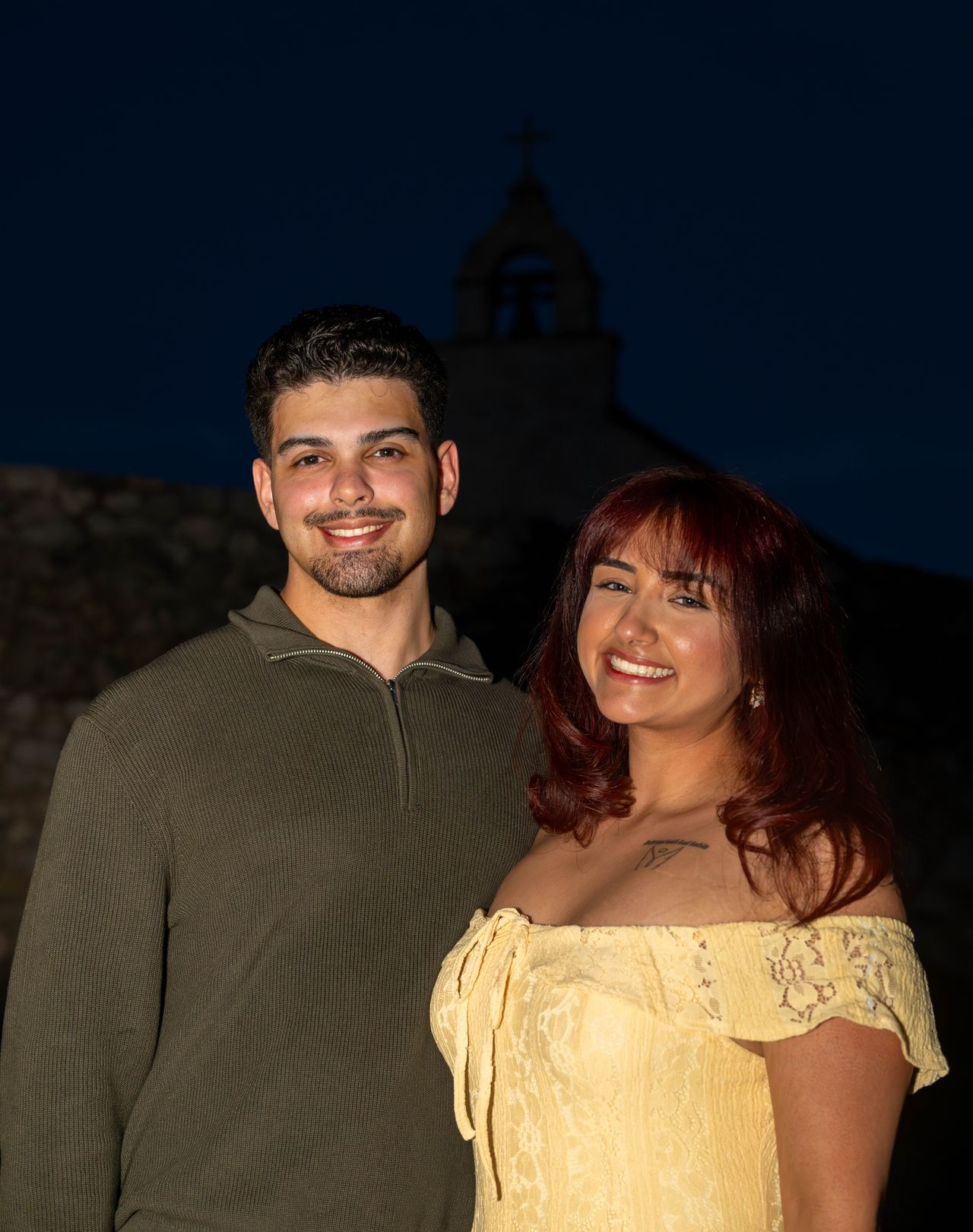 Luca and Ana smile at the camera, the chapel tower behind them