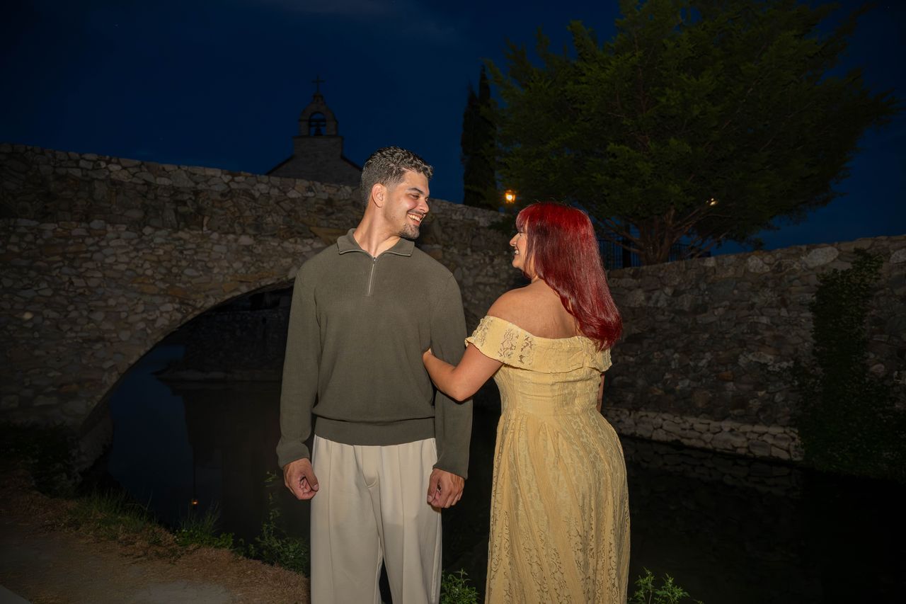 Ana holds Luca's arm beside the stone bridge at night