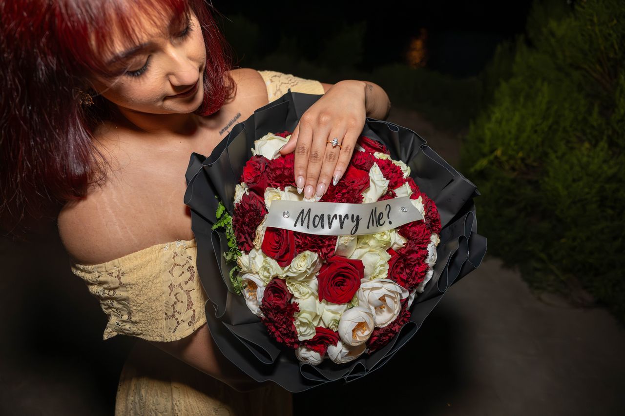 Ana rests her ring hand on the Marry Me bouquet of red and white flowers